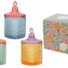 Tre scatole in vetro colorato con coperchio, disponibili in tre colori assortiti, accanto a una confezione decorata.