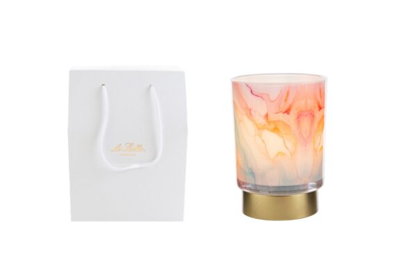 Lampada in vetro decorata con effetto acquerello multicolore, base dorata, accanto a shopper bianca con manici.